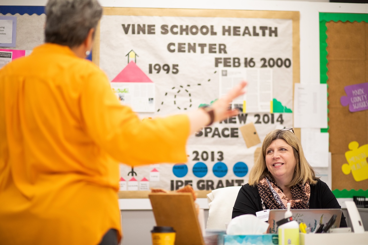 A woman wearing a black top and patterned scarf smiles while seated at a desk, engaging in conversation with another person standing in the foreground, who is wearing an orange shirt. Behind them is a bulletin board displaying a timeline of the Vine School Health Center with notable dates and milestones.