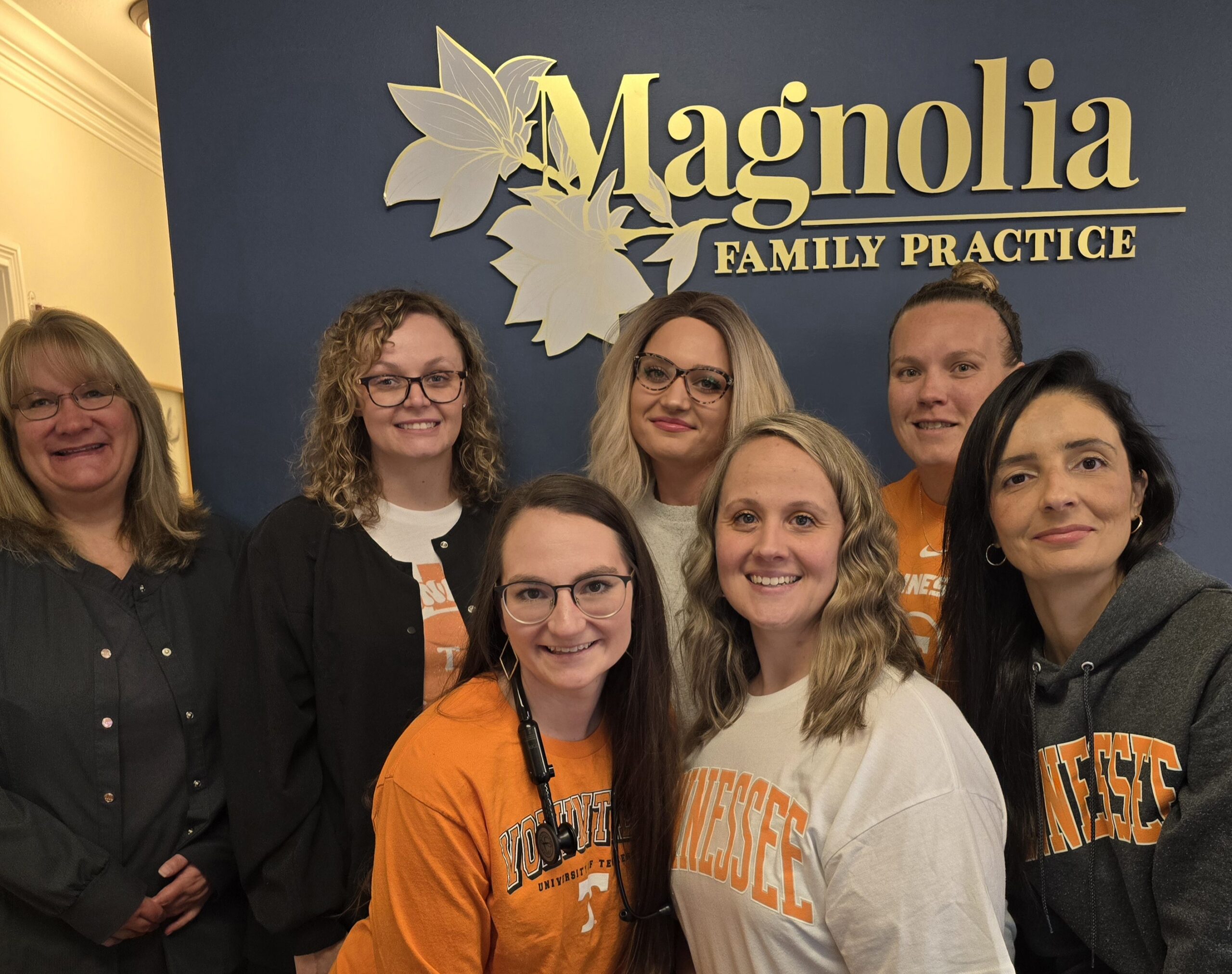 Staff at the Magnolia Family Practice stand in front of a navy blue wall dressed in UT attire.