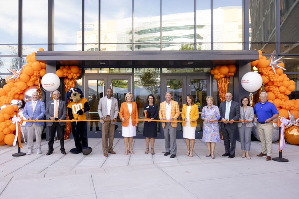 Twelve individuals stand in front of a orange ribbon with scissors to cut a ribbon to a new building seen in the background with orange balloons surrounding it.