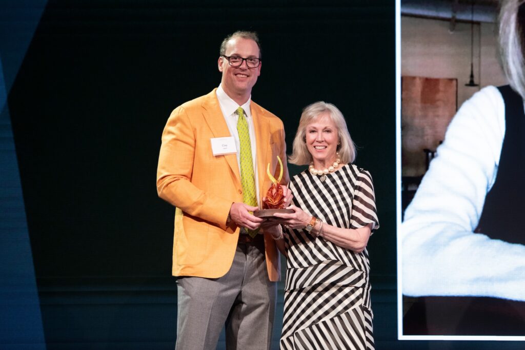 A woman is accepting an award shaped like a flame from a man. They are both looking at the camera from stage and smiling. 