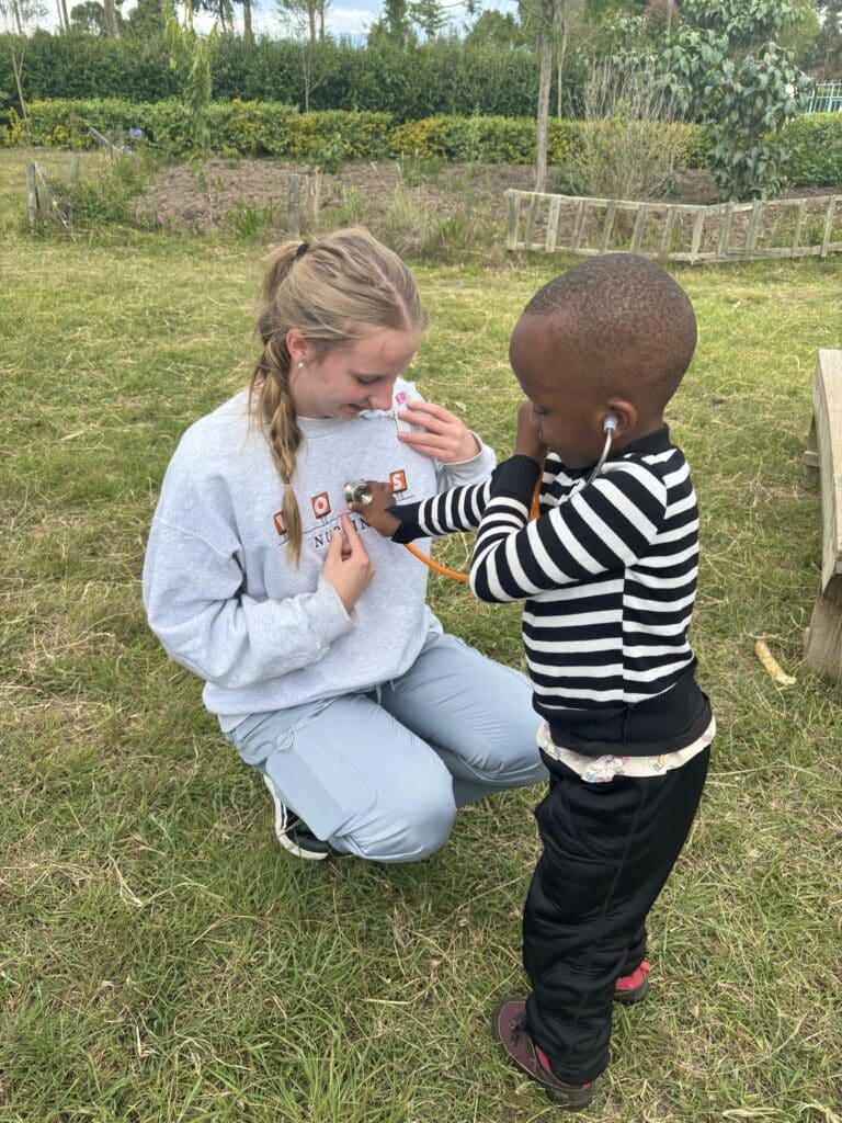 A young child is listening to the heartbeat of a nursing student with an orange stethoscope.