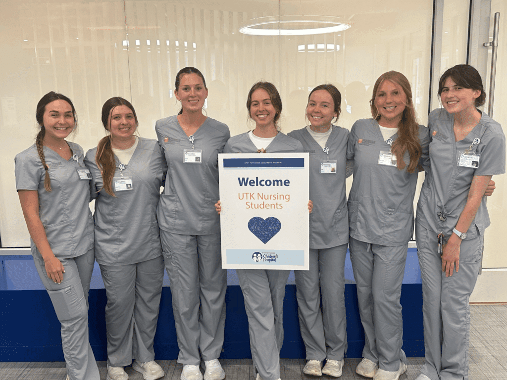 Seven nursing students are standing in front of a blue bench inside East Tennessee Children's Hospital smiling and posing while holding a sign that reads "Welcome UTK Nursing Students".